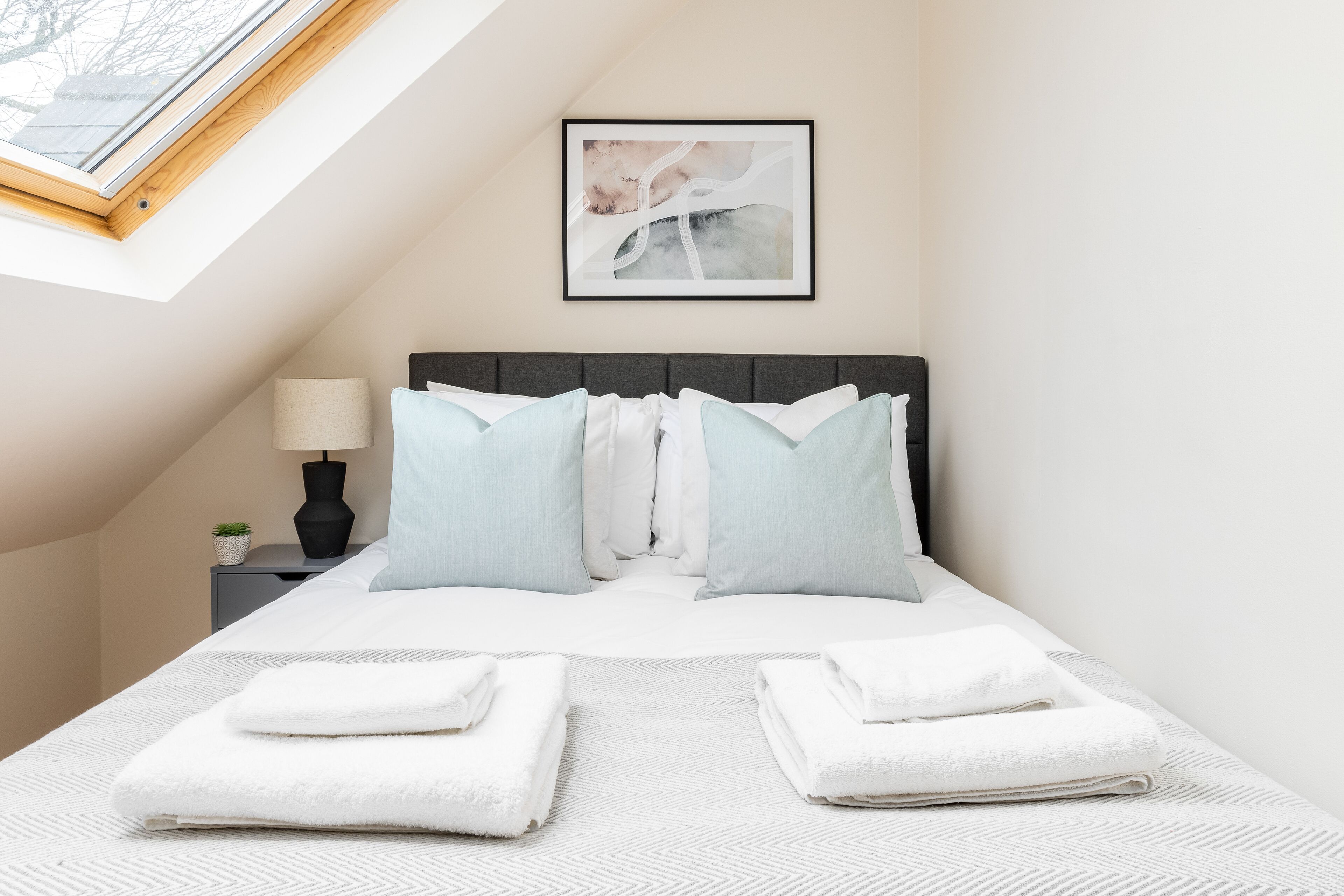Een net opgemaakt bed met witte lakens en een grijs hoofdbord, een nachtkastje met een lamp en een kleine plant, en een lijst met abstracte kunst aan de muur.