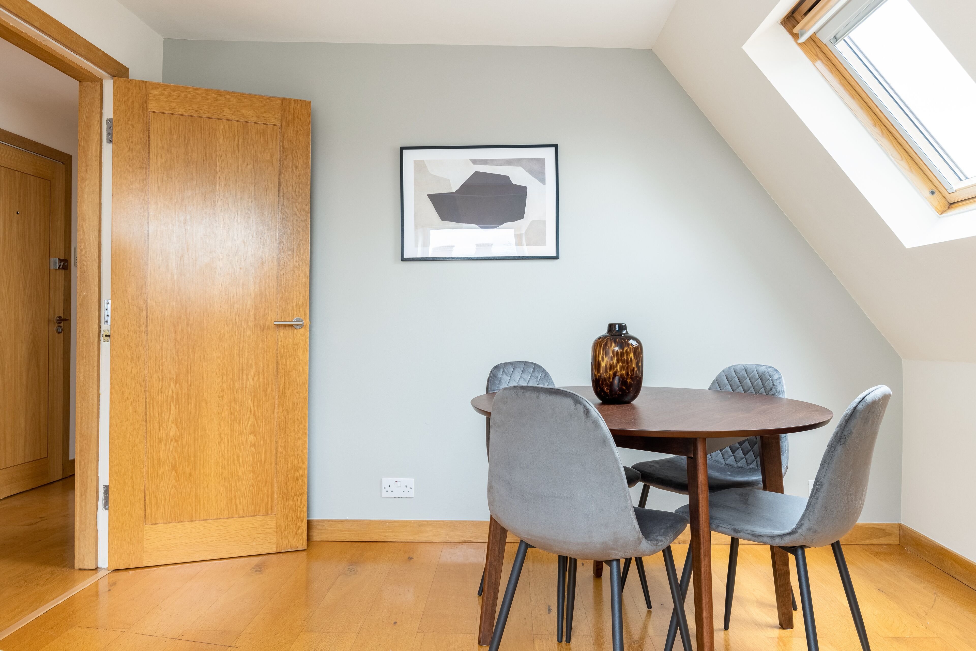A dining area with a wooden table, grey upholstered chairs, and a framed abstract artwork on the wall.