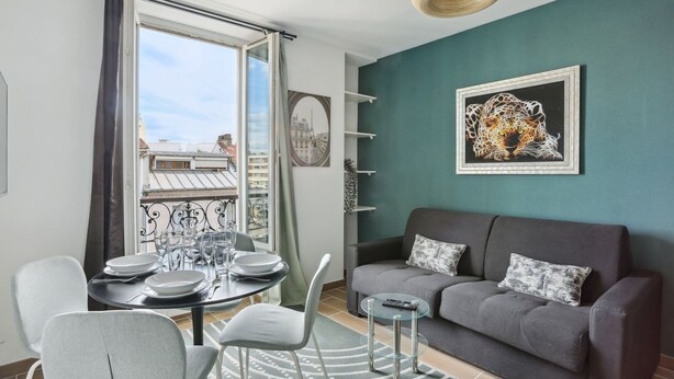 Ein modernes Wohnzimmer mit einem grauen Sofa, einem Esstisch für vier Personen und einem großen Fenster mit Vorhängen.