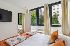 Ein Hotelzimmer mit Bett, Fernseher, Fenster mit Vorhängen und Blick auf Grünfläche.