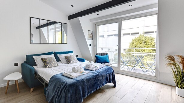 A modern bedroom with a bed, a small table, and a chair. There is a balcony with a view of the city.