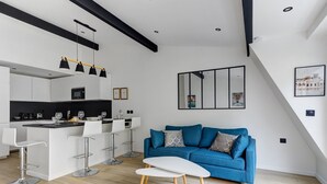 A modern kitchen with white cabinets, a black countertop, and a central island with bar stools. There is a blue sofa with patterned cushions and a white coffee table in the living area.