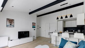 A modern living room with a flat-screen TV, white furniture, a blue sofa, and a kitchen area with bar stools.