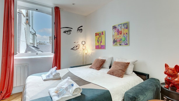 Een hotelkamer met een bed, kussens, een raam met rode gordijnen en een kunstwerk aan de muur.