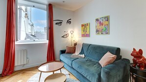 Een woonkamer met een blauwe bank, een kleine ronde tafel, rode gordijnen en een kunstwerk aan de muur.