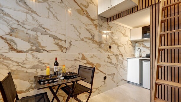 A dining area with a marble wall, a table set with plates, glasses, and a bottle, and a kitchen in the background. - 366 Suite Legendre - Superb apartment in Paris (Paris 17 Batignolles-Monceau)