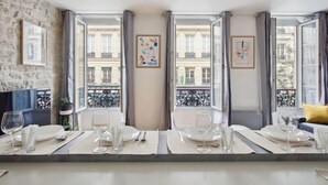 A dining area with a table set for four, large windows with curtains, and a view of a building with balconies. - 626 Suite Rambuteau - Superb apartment in Paris (Paris 04 Hôtel-de-Ville)