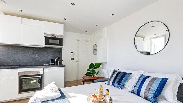 Una habitación de hotel moderna con kitchenette, una cama con almohadillas a rayas, una mesita de noche con una cesta de pan y bebidas, y un espejo grande en la pared.