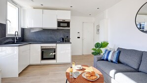 Una cocina moderna con armarios blancos, un horno empotrado y un microondas. Hay una mesa de comedor con una fuente de cruasanes y un vaso de agua. Un sofá gris se encuentra en la sala de estar, con un espejo grande en la pared.