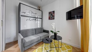 A modern living room with a grey sofa, a black TV mounted on the wall, a small table with a plant, and a yellow patterned rug. - Suite CD : Superb Parisian apartment (Paris)