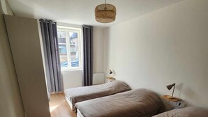 Quarto pequeno e moderno, com duas camas, ventoinha no teto, janela com cortinas e uma mesinha com abajur.