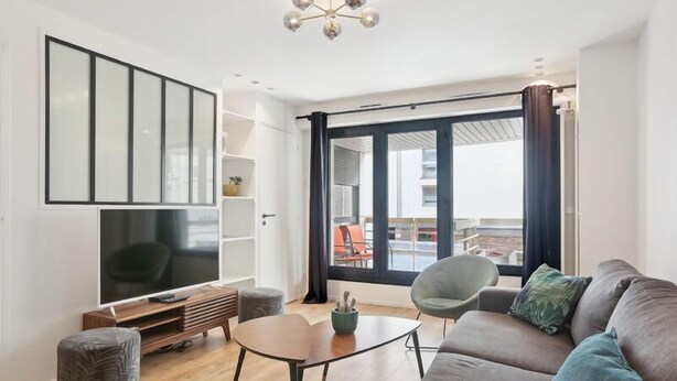 A modern living room with a large flat-screen TV, a sofa, a coffee table, and a chair. There is a sliding glass door leading to an outdoor area with a view of the city. - 174 Suite Grenelle - Marvelous apartment in Paris (Paris 15 Vaugirard)