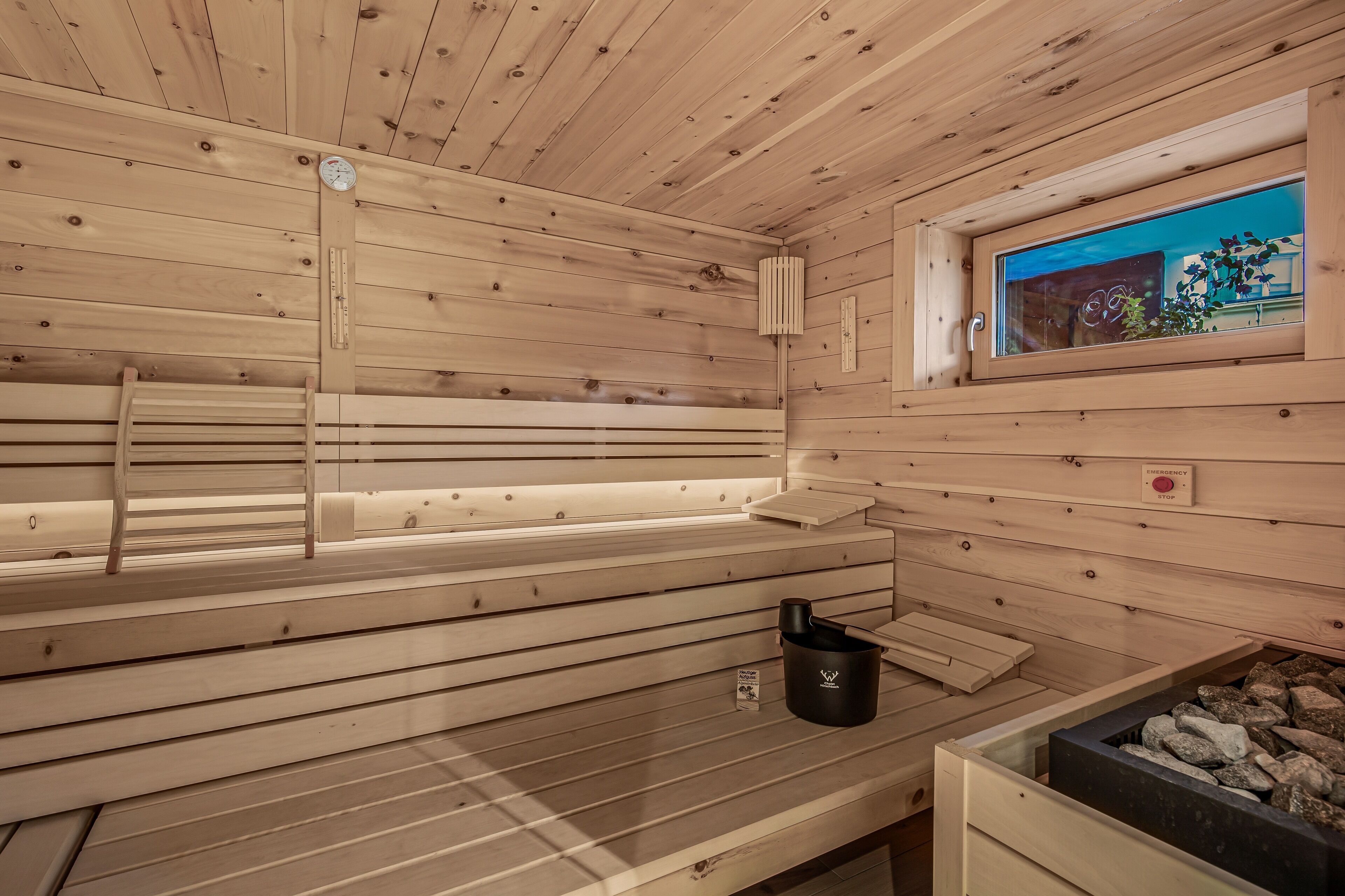 A sauna with wooden benches, a thermometer, a window with a view, and a black container.