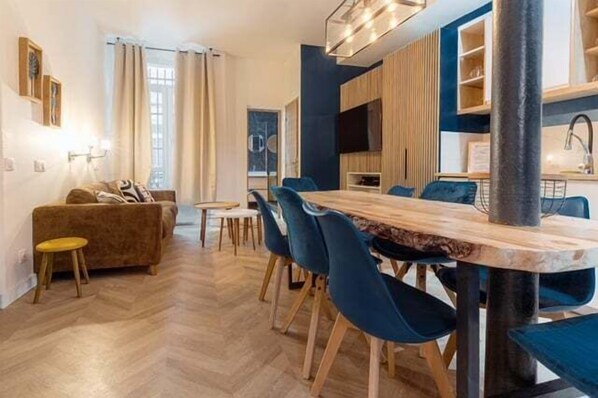 A modern living room with a wooden dining table and blue chairs, a sofa, and a small stool.