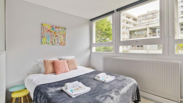 Ett modernt hotellrum med en stor säng, ett fönster med utsikt över byggnaden, ett element och ett färgstarkt konstverk på väggen.