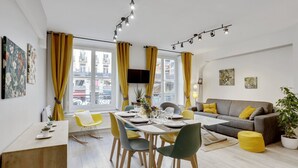 A modern living room with a dining area, large windows with yellow curtains, a grey sofa with yellow cushions, and a flat-screen TV mounted on the wall. - 215 Suite Esprit Nature - Superb apartment (Paris 02 Bourse)