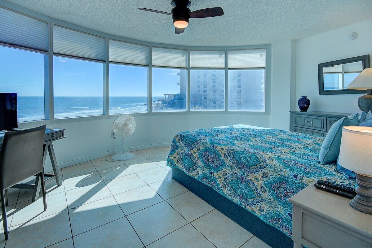 Un dormitorio con una cama grande, un escritorio, una silla, un ventilador de techo y vista al océano.