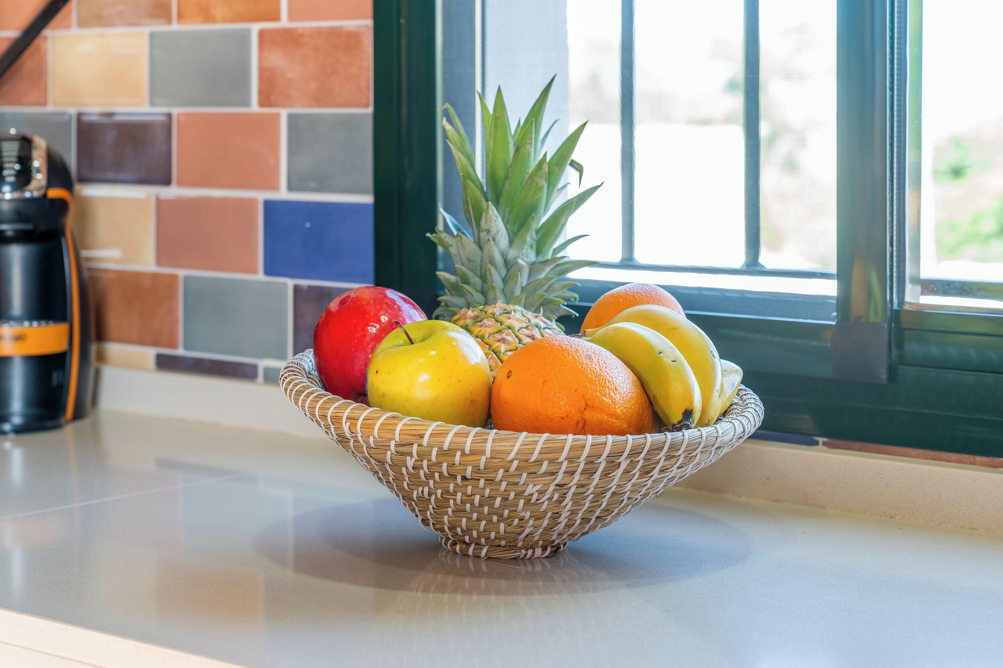 Un bol de mimbre con fruta sobre una encimera de cocina, con un salpicadero de azulejos coloridos y una ventana al fondo.