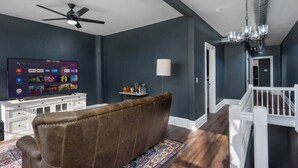 Een woonkamer met een donkerblauwe wand, een bruine leren bank, een flatscreen televisie, een plafondventilator en een kroonluchter.