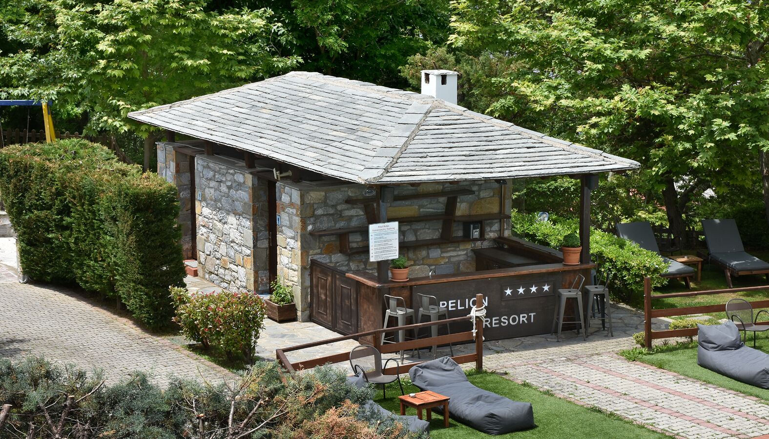 Een klein stenen gebouw met een bord waarop ‘PELION RESORT’ staat, en een terras met zitplaatsen buiten.