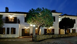 A two-story building with a stone facade, multiple windows with shutters, and a central tree. - Hotel Pelion Resort (Volos)