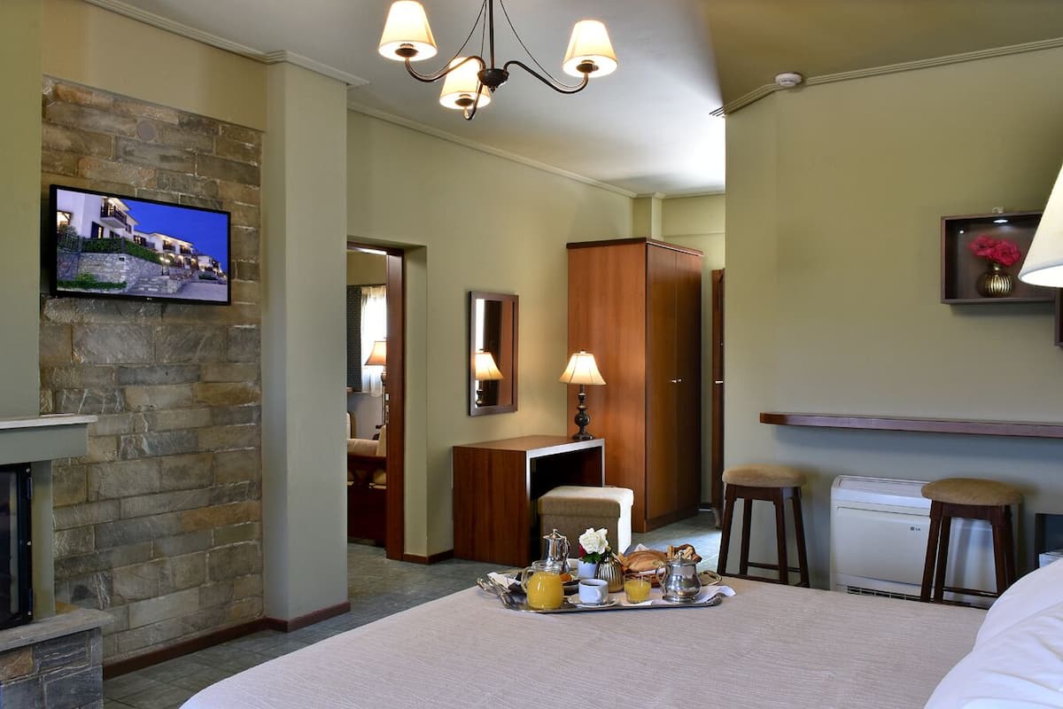 A hotel room with a stone fireplace, a television mounted on the wall, a dining area with a tray of breakfast items, and a bed with white linens.