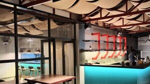 Et moderne restaurantmiljø med træborde, sorte stole og en blå bardisk.