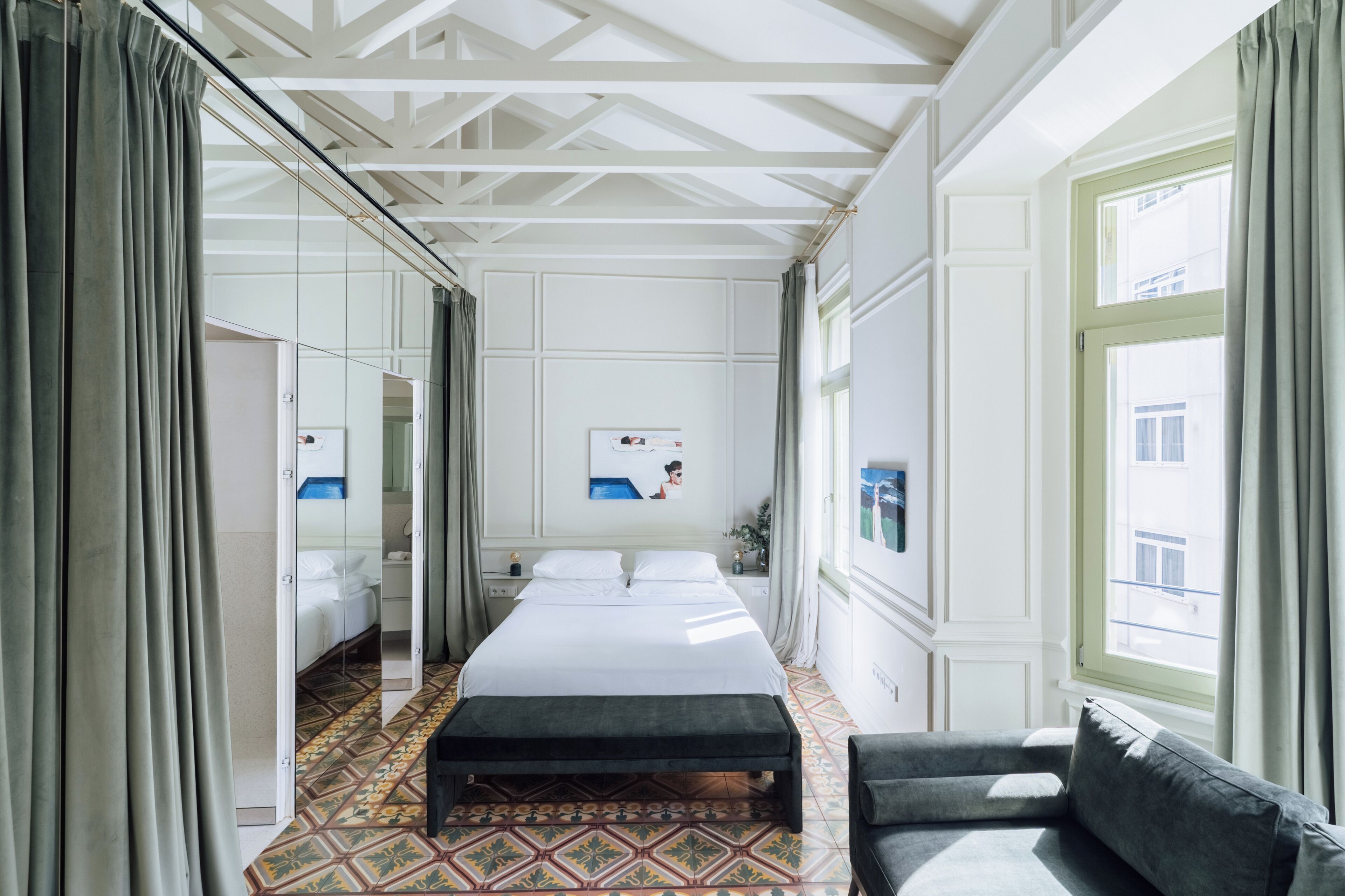 Habitación de hotel con una cama grande, un banco, un sofá, un espejo y una ventana con cortinas.