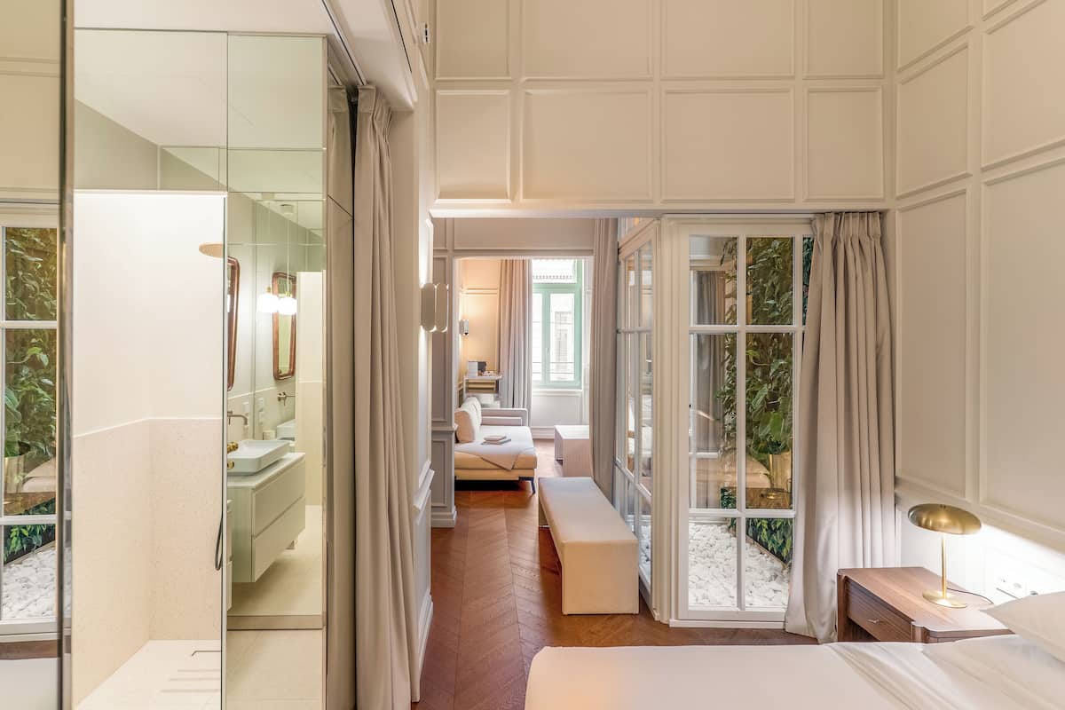 Habitación de hotel con un espejo grande, una cama, un banco, una mesita de noche y una ventana con cortinas.