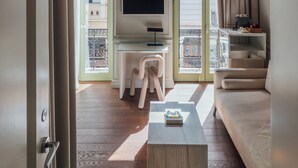Una habitación con televisión sobre un soporte blanco, un sofá, una mesa de centro y un balcón con vistas a edificios.