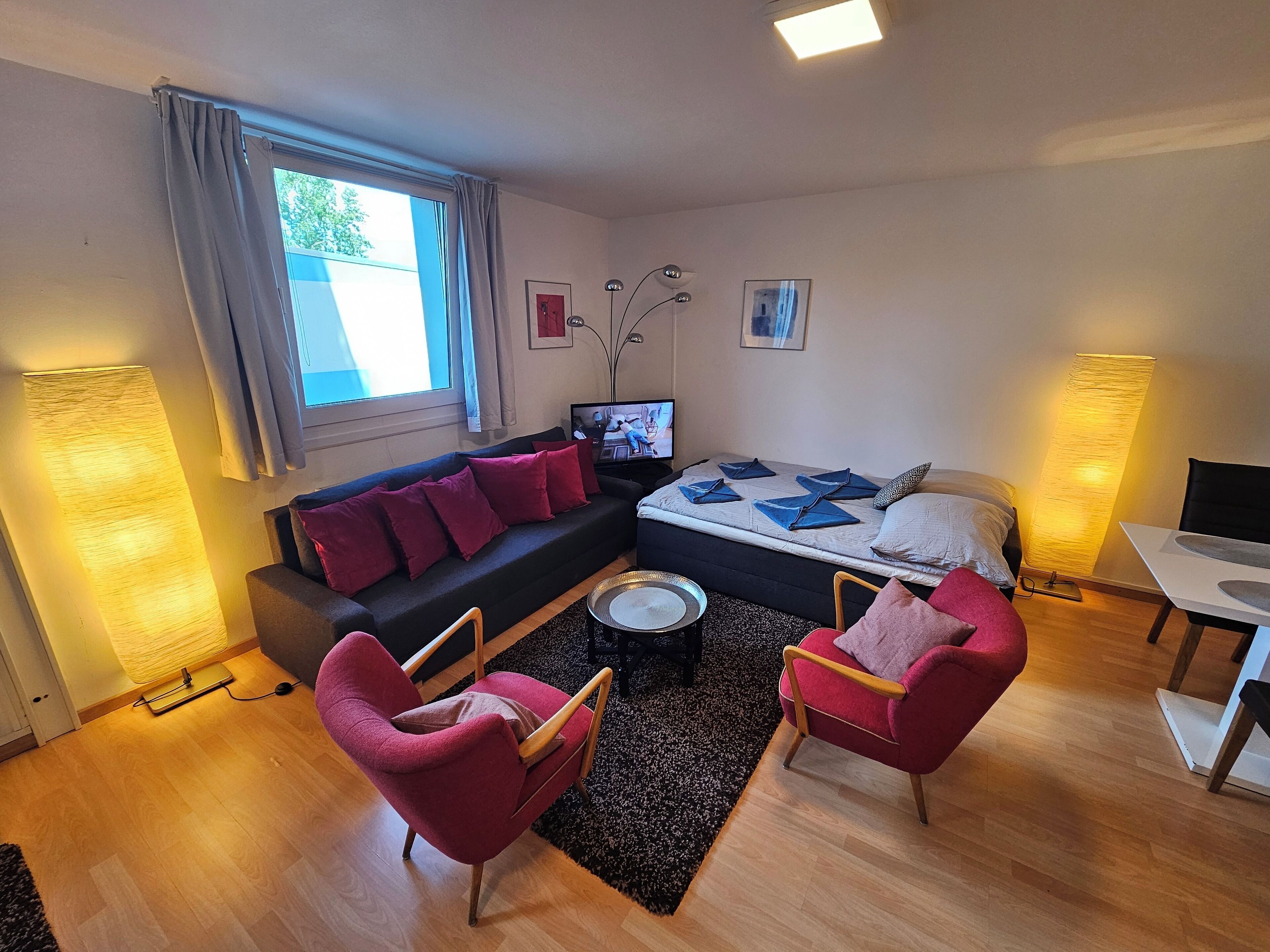 Ein modernes Wohnzimmer mit einer Couch, zwei Sesseln, einem Couchtisch, einem Bett mit Kissen, einem Fernseher und einer Lampe.