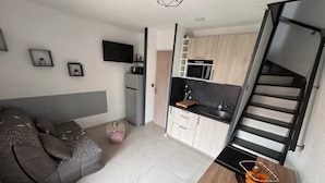 A compact living space with a sofa, a small round table, a kitchen area with a microwave and a sink, and a TV mounted on the wall.