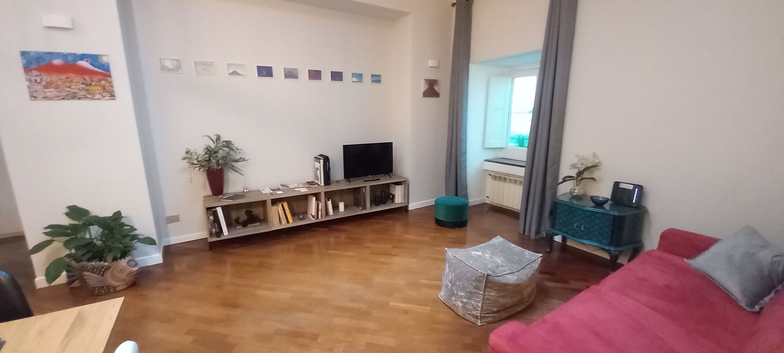 Una sala de estar con piso de madera, un sofá rojo, un televisor sobre un mueble bajo, una estantería y una planta en maceta.