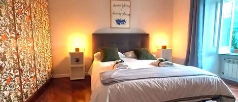 Une chambre à coucher avec un lit, deux tables de chevet, un mur orné d’un motif floral et une fenêtre avec des rideaux.