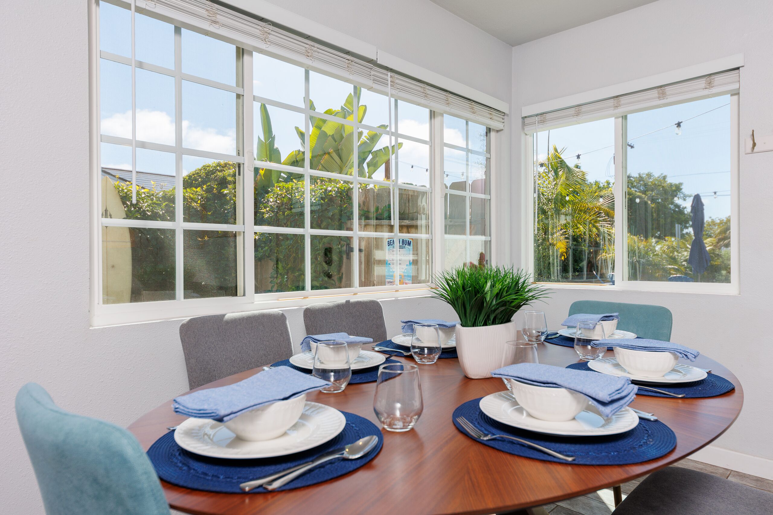 A dining area with a wooden table set for four, surrounded by chairs, with a view of greenery outside.