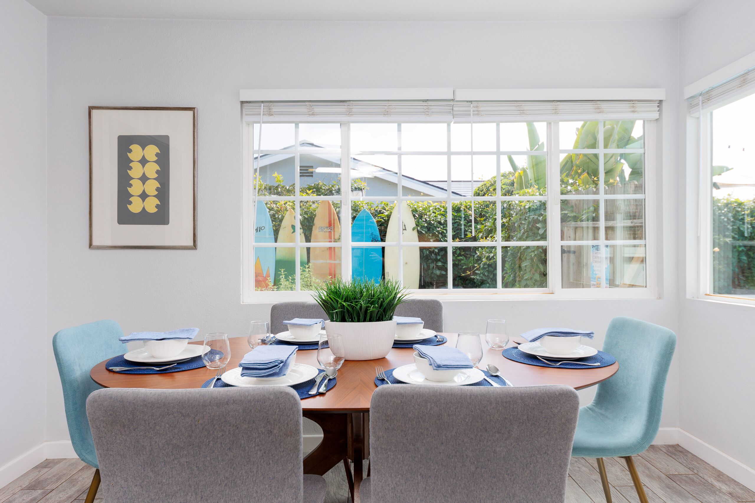 Un comedor con una mesa de madera dispuesta para cuatro personas, rodeada de sillas grises. Hay una planta en maceta sobre la mesa y un cuadro enmarcado en la pared.