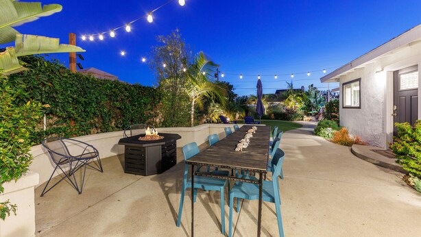 Un patio al aire libre con una mesa larga de comedor, sillas y luces colgantes.