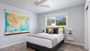 Un dormitorio con cama, ventilador de techo, ventana con persianas, un mapa en la pared y una mesita de noche con lámpara.