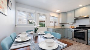 Un comedor con una mesa de madera dispuesta para cuatro personas, rodeado de sillas azules. Se ve una cocina de fondo con armarios azul claro y un horno de acero inoxidable.