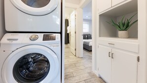 Un área de lavandería con lavadora de carga frontal y secadora, un pasillo que conduce a un dormitorio y un armario blanco con una planta.