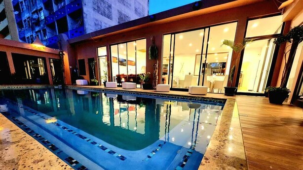 A swimming pool with a tiled deck, adjacent to a building with large windows and indoor seating. - Villa das Arabias (Maputo)