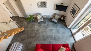 Un vaste espace de vie comprenant un canapé rouge, deux fauteuils, une petite table et une plante en pot. On aperçoit à l’arrière-plan un escalier avec des marches en bois et une rampe en métal.