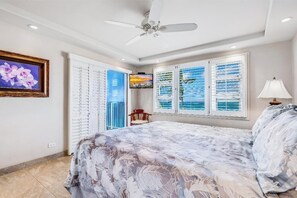 Une chambre à coucher avec un lit, un ventilateur de plafond, une fenêtre avec des stores, un tableau encadré au mur et une chaise.