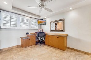 Une pièce comprenant un bureau, une chaise, un ventilateur de plafond et une fenêtre avec des stores.