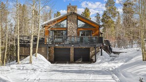 A log cabin with a stone chimney, a balcony with chairs, and a snow-covered driveway.