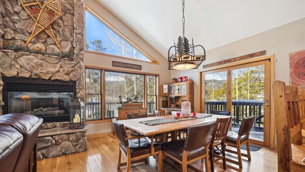 A cozy living room with a stone fireplace, a dining table with chairs, and a large window overlooking a deck.