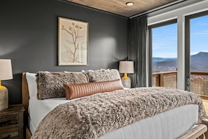 A bedroom with a large bed, two bedside lamps, a framed botanical print on the wall, and a view of the landscape through large windows.