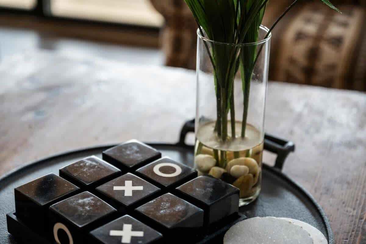 Un tablero de juego negro con símbolos blancos, un jarrón de vidrio con plantas verdes y un pequeño recipiente con piedras sobre una superficie de madera.