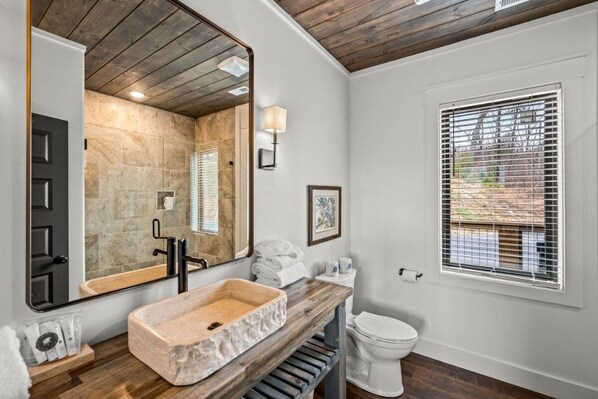 A bathroom with a stone sink, wooden vanity, a large mirror, a toilet, and a window with blinds.