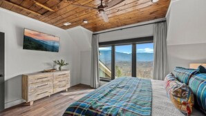 A bedroom with a wooden ceiling, a bed with plaid bedding, a dresser, and a large window with a mountain view.